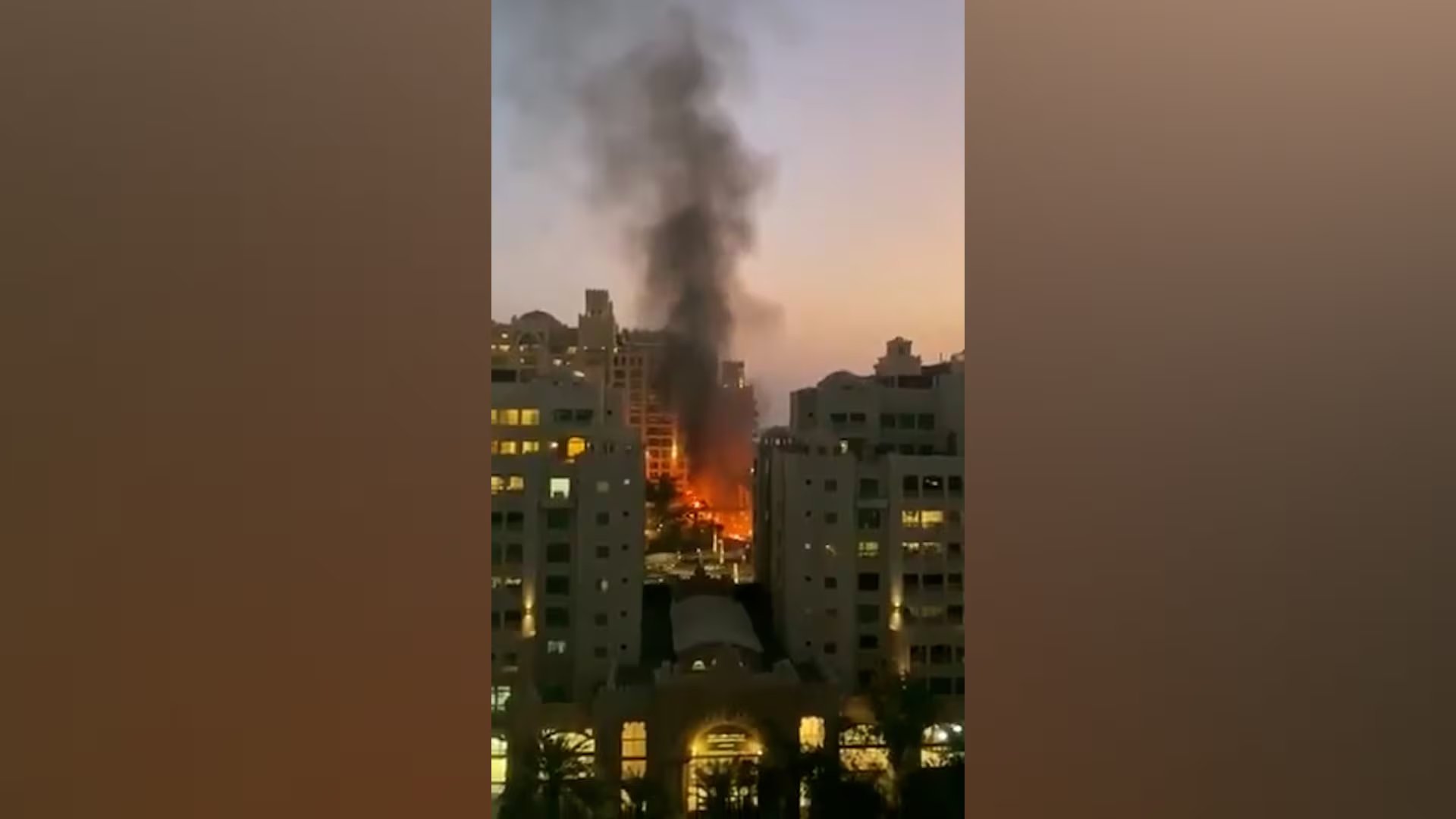 Un gran incendio consume un edificio alto de apartamentos o un rascacielos con intensas llamas y una gruesa columna de humo negro que se alza contra el cielo del atardecer, visto entre otras edificaciones.