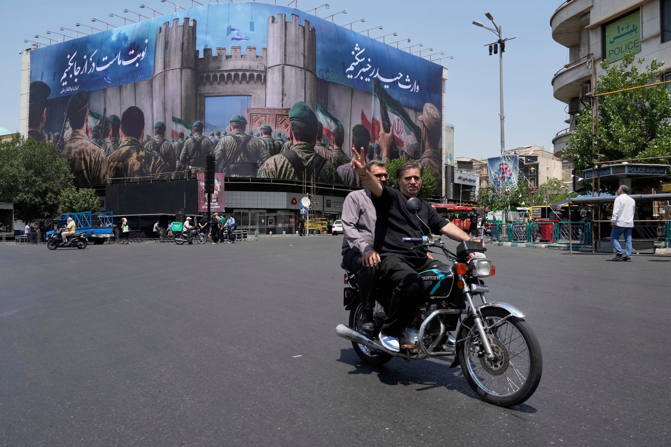 Un hombre hace el signo de la victoria desde su motocicleta tras pasar frente a una manta que muestra a soldados iraníes que se encaminan a atacar territorio israelí, en la Plaza de la Revolución Islámica, el sábado 14 de junio de 2025, en Teherán, Irán (AP Foto/Vahid Salemi)