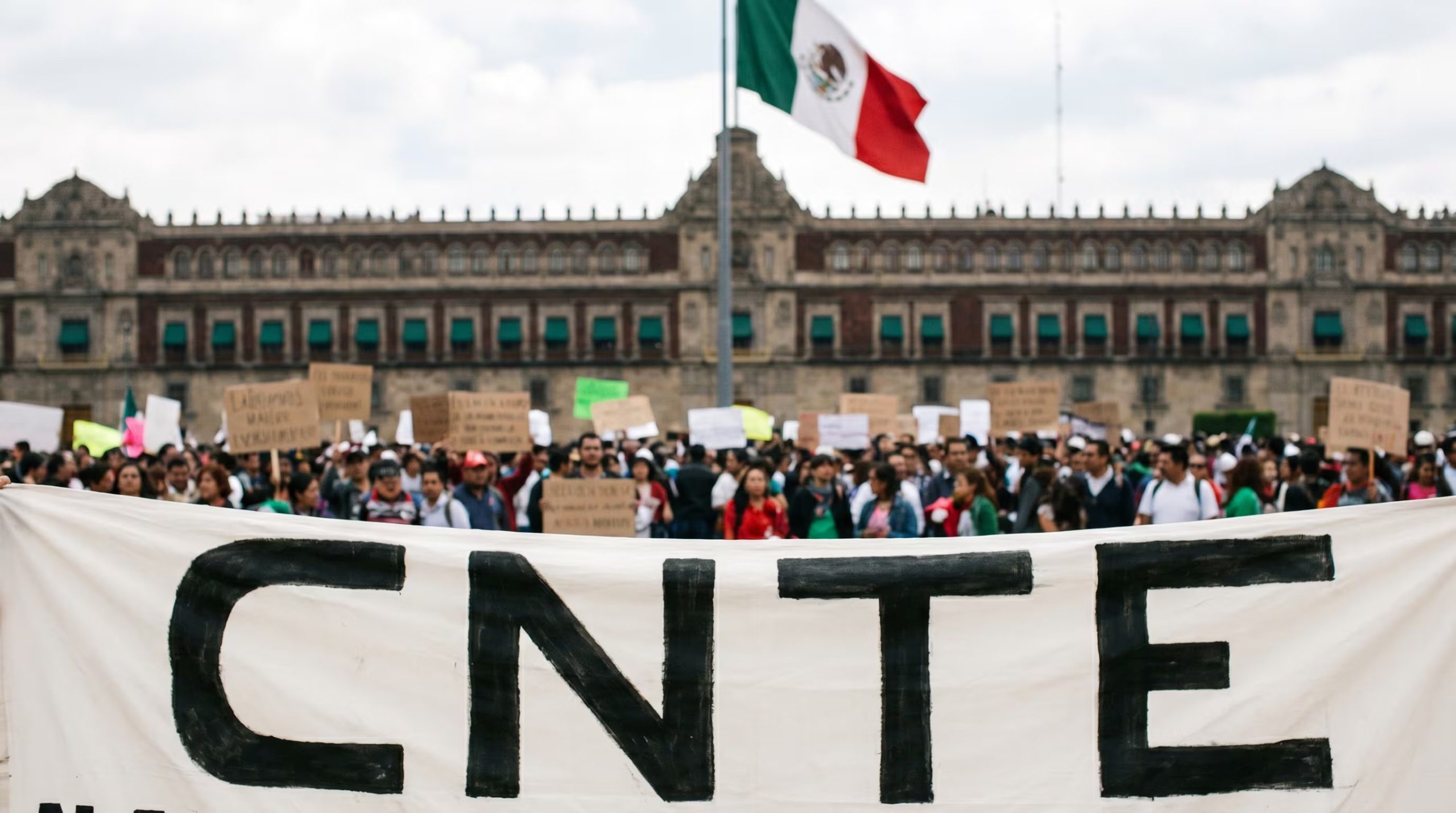Una multitud de maestros, algunos con cartulinas, se congrega frente al Palacio Nacional de México, con una pancarta de la CNTE en primer plano, durante una movilización tensa