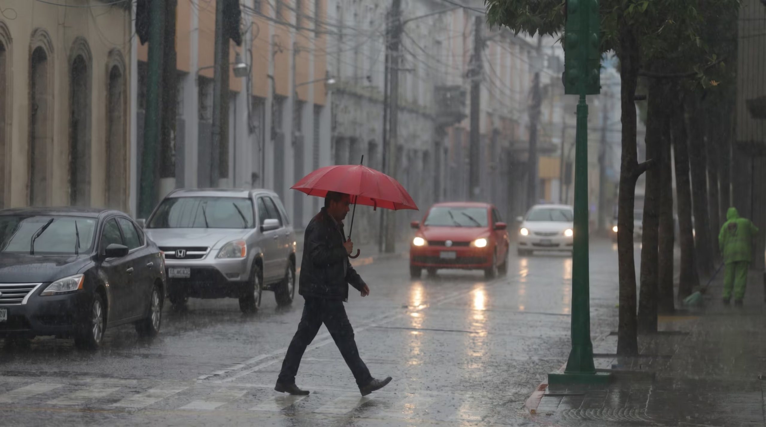 Un hombre camina con un paraguas rojo bajo una lluvia intensa, cruzando una calle mojada mientras varios vehículos esperan el paso, reflejando el impacto del temporal en el tránsito urbano. (Imagen Ilustrativa Infobae)