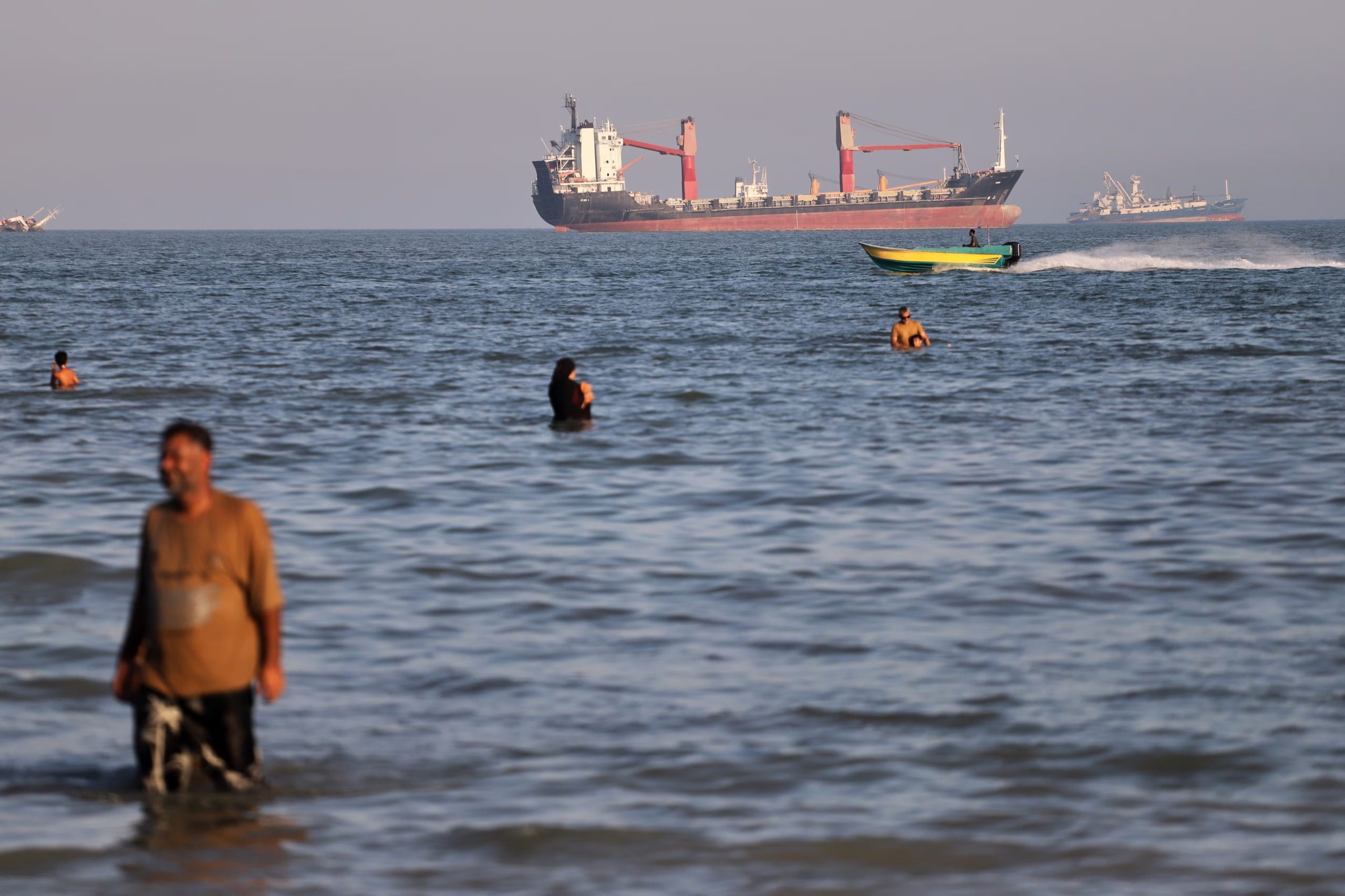 Residentes nadan en las aguas del estrecho de Ormuz mientras una pequeña lancha pasa junto a cargueros y otros buques comerciales en alta mar, cerca de Bandar Abbas, Irán, el 17 de junio de 2026 (Amirhosein Khorgooi/ISNA vía AP)