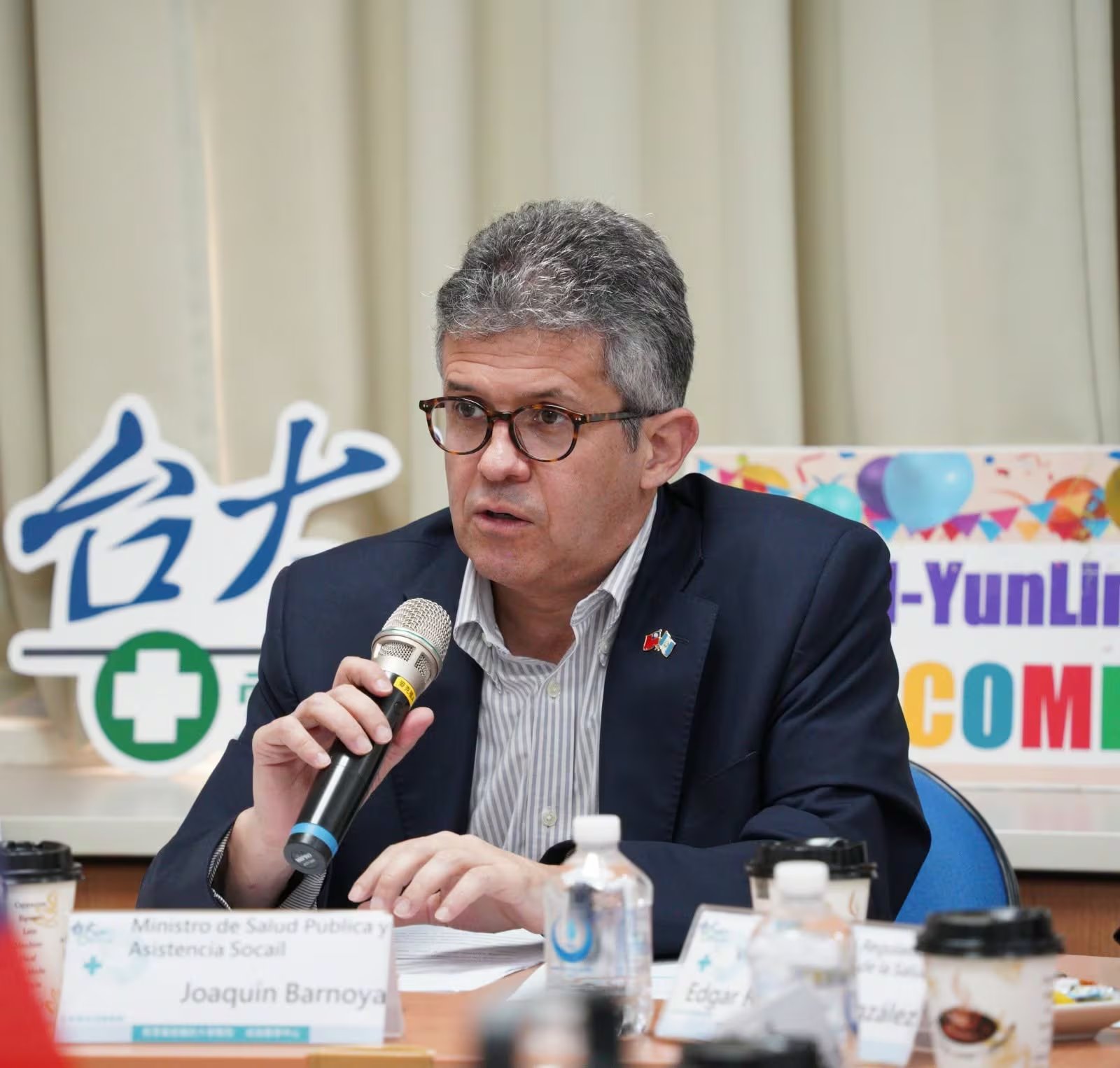 El Ministro de Salud Pública y Asistencia Social de Guatemala, Joaquín Barnoya, interviene en un evento público con un micrófono en la mano, frente a una mesa con bebidas. (Ministerio de Salud Pública y Asistencia Social de Guatemala)