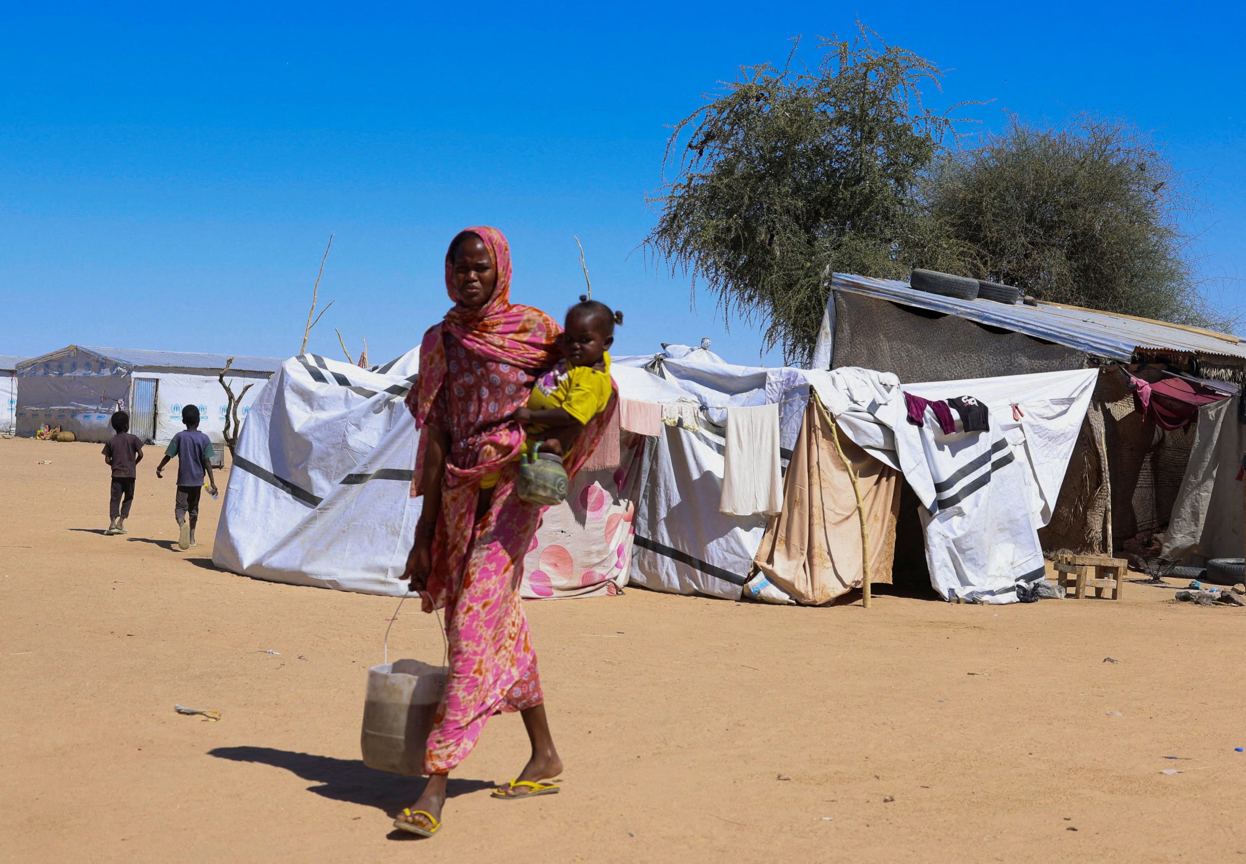 Una mujer refugiada sudanesa de Al-Fashir carga a un niño y agua, con tiendas de campaña al fondo, en el campo de refugiados de tránsito de Tine en medio del conflicto entre las Fuerzas paramilitares de Apoyo Rápido (RSF) y el ejército sudanés, en el este de Chad (REUTERS/Amr Abdallah Dalsh)