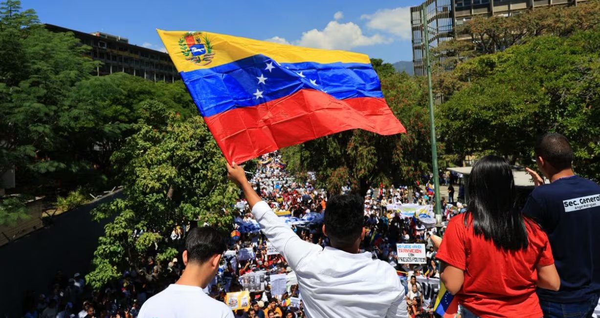 Un hombre levanta orgullosamente la bandera de Venezuela sobre una multitud de personas reunidas en una manifestación cívica ((Comando Con Venezuela))