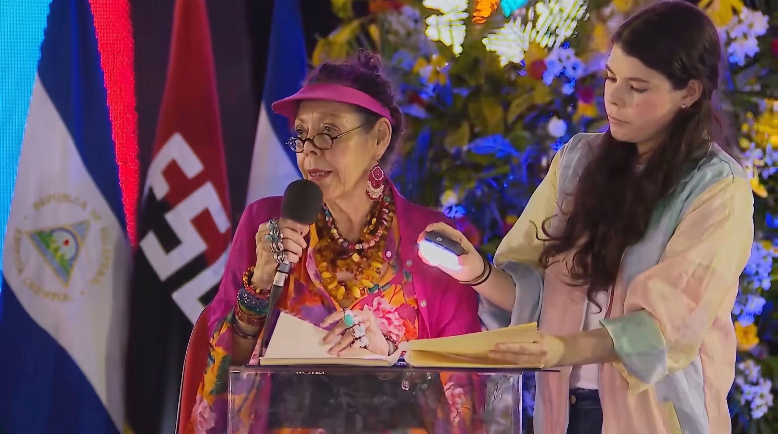 Rosario Murillo, vicepresidenta de Nicaragua, habla en un acto público mientras una asistente ilumina sus documentos sobre un atril transparente. (Imagen Ilustrativa Infobae)